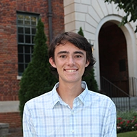 A young man smiling in front of a brick building and neatly manicured greenery.