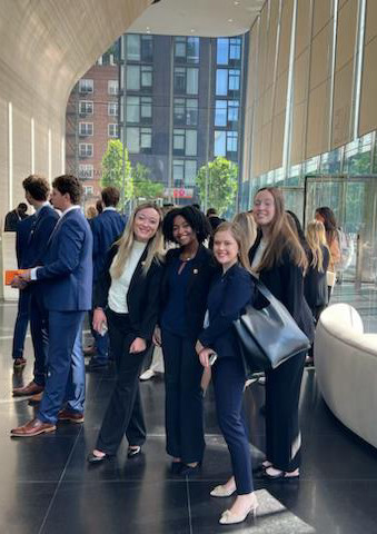 A group of four young professionals posing together in a modern office lobby, dressed in business attire.