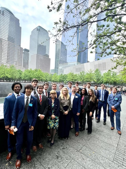 Group photo of young men and women in formal attire gathered outdoors at a memorial site in a city, with skyscrapers and greenery in the background.