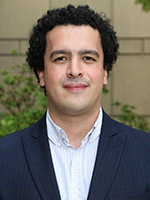Portrait of a young man with curly hair wearing a blazer and a collared shirt, standing outdoors with greenery in the background.