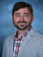 Professional headshot of a smiling man with a beard, wearing a plaid shirt and light gray blazer.