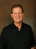 A smiling man in a black polo shirt against a neutral background.