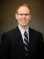 Professional headshot of a man in a suit and tie, smiling against a neutral background.