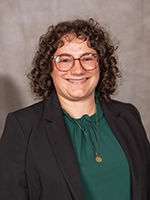 Smiling person with curly hair and glasses wearing a dark blazer over a green blouse, posed against a neutral studio backdrop.