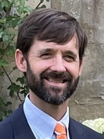 Portrait of a man with a beard and a warm smile, wearing a suit and an orange tie, against a natural background.