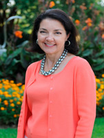 A professional portrait of a woman smiling, wearing an orange cardigan and a statement necklace, set against a backdrop of vibrant flowers.