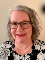 A smiling woman with glasses and wavy hair wearing a black and white patterned top and a chunky necklace.