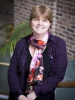 A smiling woman with short brown hair, wearing a purple jacket and a colorful scarf, poses in a well-lit indoor setting.