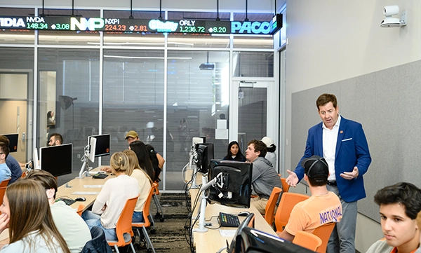 A classroom setting where students are engaged at computer stations, with an instructor speaking to a group. An electronic ticker displaying stock prices is visible above.