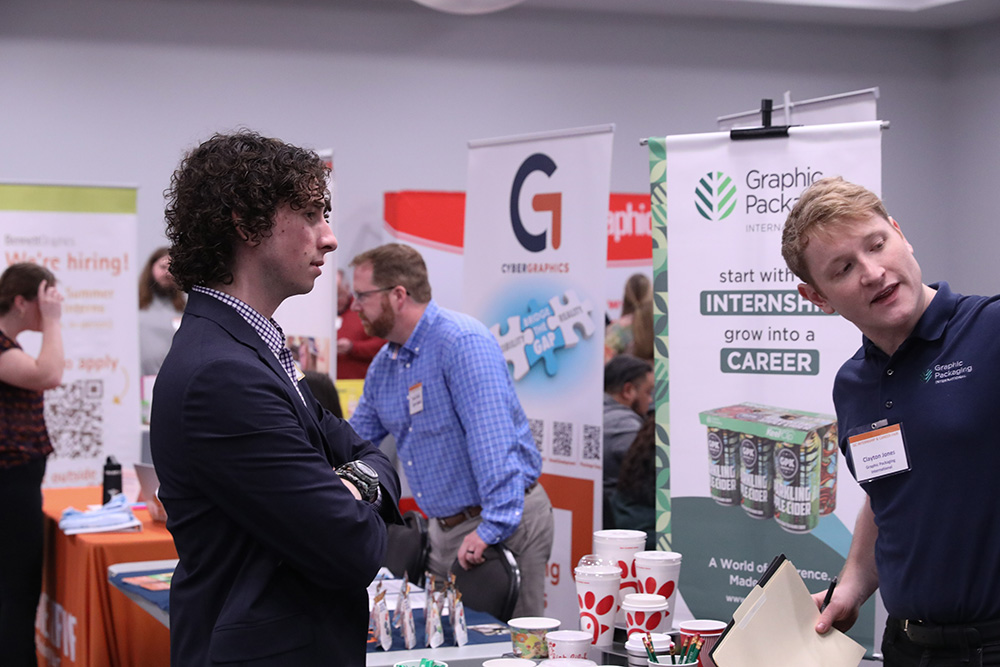 A woman engages with a visitor at a trade show booth, showcasing promotional materials and branded products.