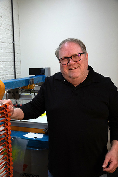 A smiling man in a black shirt stands next to a machine in a workshop, holding orange circular objects.