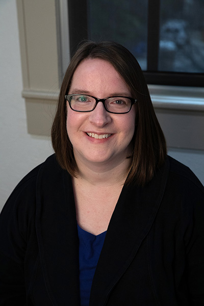 A professional portrait of a woman with glasses and shoulder-length hair, smiling warmly while seated indoors.