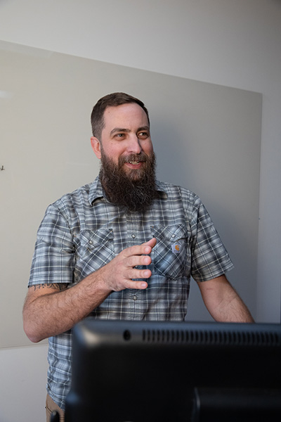 A man with a beard wearing a plaid shirt stands in front of a computer, gesturing as he speaks.