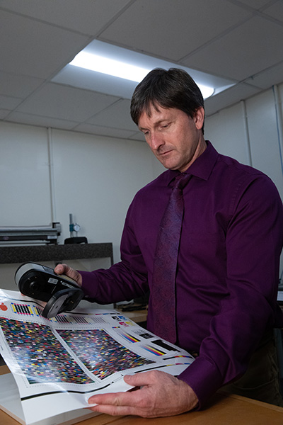 A professional man in a purple shirt and patterned tie examines a color printout with a color measurement device in a well-lit office.