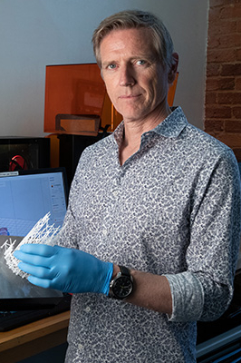 A person wearing blue gloves holds a 3D printed object while standing in a workspace with computer equipment in the background.