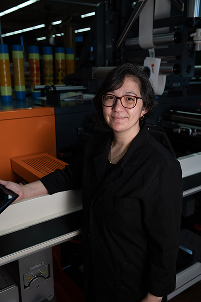 A woman in black attire stands next to a printing machine, holding a tablet, in a brightly lit industrial setting.