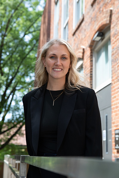 A smiling woman in a black blazer stands outdoors in front of a brick building, with trees in the background.