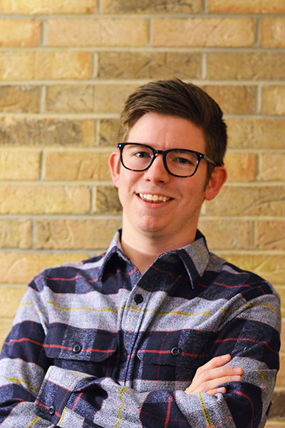 A smiling young man with glasses, wearing a plaid shirt, poses confidently against a brick wall.
