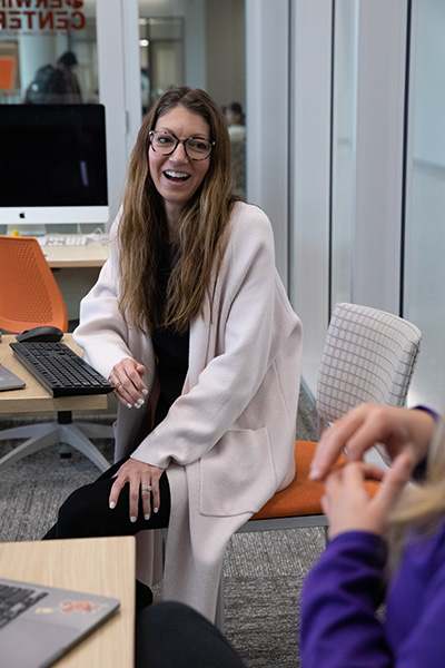 A smiling woman with long hair wearing glasses, sitting on an orange chair in a modern office environment.
