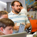 A man with a beard wearing a striped shirt engages with a group in a collaborative workspace.