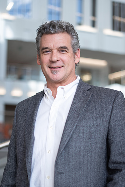 Professional portrait of a man with curly hair wearing a gray blazer and white shirt, smiling in a modern building setting.