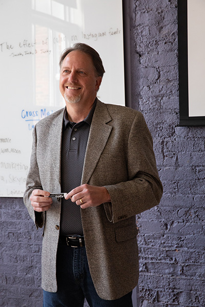 A smiling man in a gray blazer stands in front of a whiteboard, holding a pair of glasses.