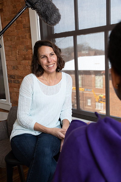 A woman with curly hair smiling while seated in an interview setting, engaged in conversation with another person.