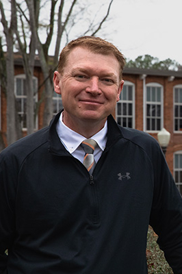 A smiling man in a black zip-up jacket and tie stands outdoors in front of a brick building, embodying a friendly and approachable demeanor.