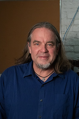 Portrait of a man with long hair and a beard, wearing a blue shirt, smiling at the camera.