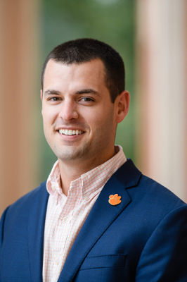 A smiling man in a navy suit jacket with a light pink shirt, featuring a Clemson University paw print lapel pin.