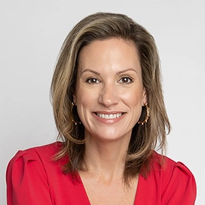 Professional headshot of a smiling woman with shoulder-length hair, wearing a red blouse and gold hoop earrings.