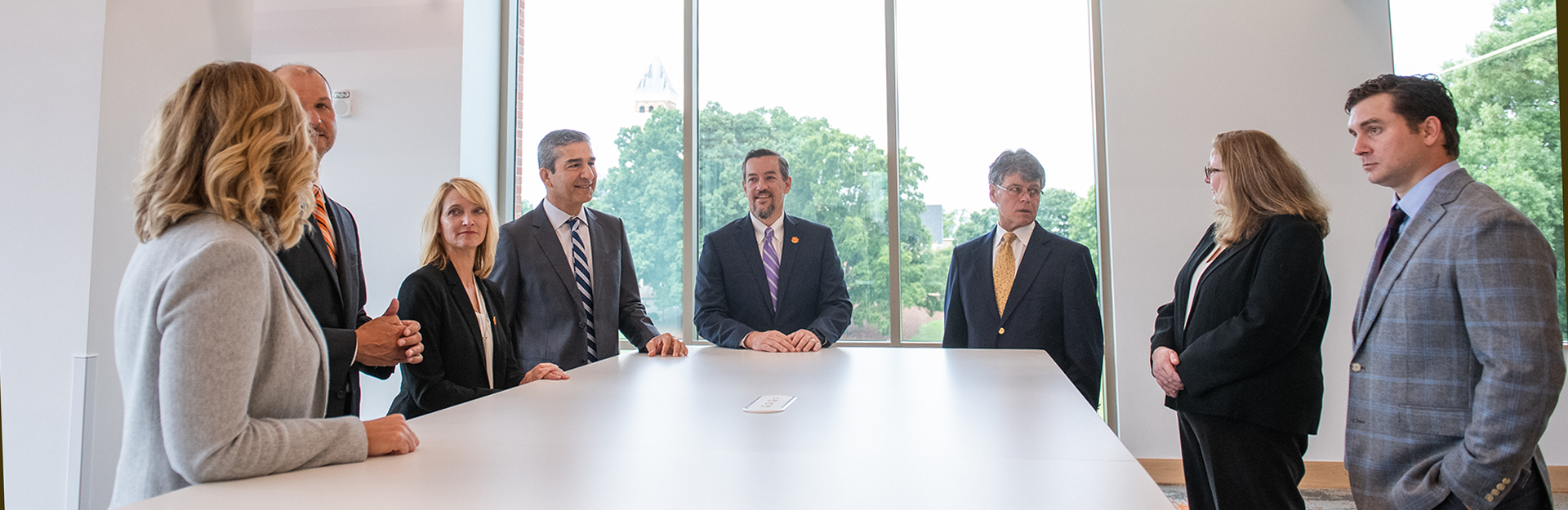 Faculty standing in conference room with Tillman Hall in the background of the window behind them.