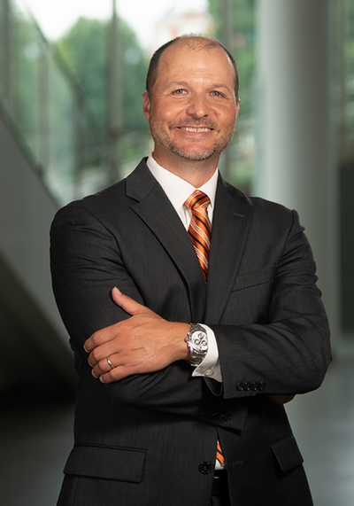 A professional portrait of a man in a suit with an orange tie, smiling confidently with arms crossed.