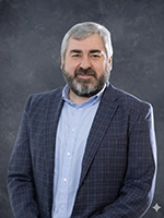 Professional portrait of a man with a beard, wearing a blue suit jacket and a light blue shirt, posing against a gray background.
