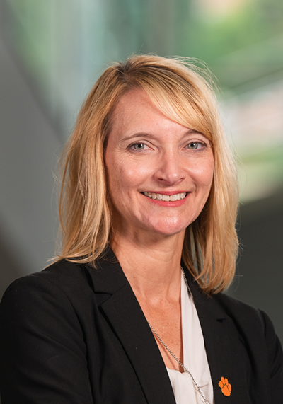 Portrait of a smiling woman with blonde hair, wearing a black blazer and a Clemson University paw print pin.