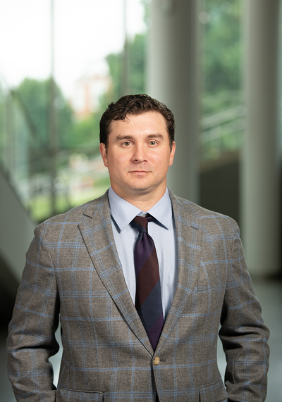 Professional portrait of a man in a plaid blazer and tie, standing in a modern building with large windows in the background.