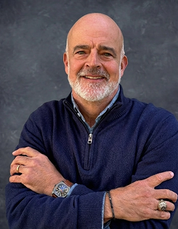 A smiling man with a beard, wearing a navy sweater, poses for a portrait with crossed arms against a textured gray background.