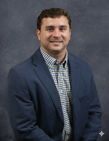 Professional portrait of a man in a blue blazer and checkered shirt, smiling against a gray background.