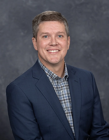 Professional headshot of a man wearing a blue blazer and plaid shirt, smiling against a gray background.