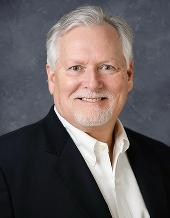 Professional headshot of a man with gray hair, smiling confidently while wearing a dark blazer over a white shirt.