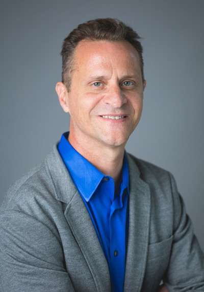 Headshot of a smiling man in a gray blazer over a blue shirt, set against a neutral background.