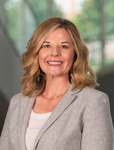 Professional portrait of a woman with shoulder-length blonde hair, wearing a grey blazer and smiling confidently.