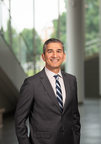 Professional headshot of a man in a suit, smiling in a modern architectural setting with large windows.