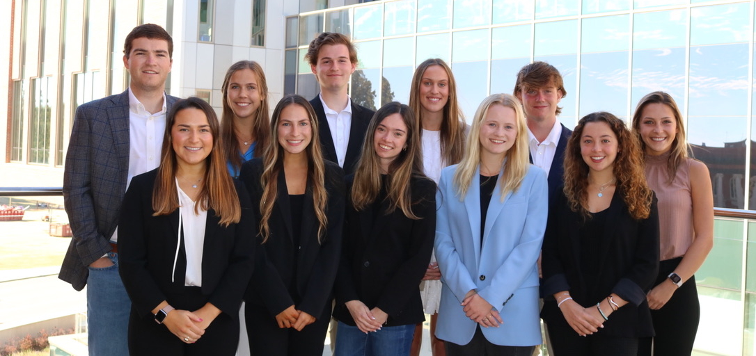 A group photo of eleven individuals, dressed in business attire, standing in front of a modern building with a reflective glass facade.