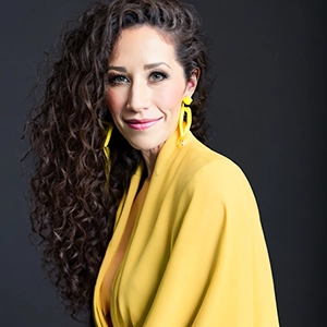 A woman with curly hair wearing a vibrant yellow outfit and large yellow earrings, smiling against a dark background.