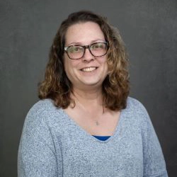 A smiling woman with curly hair wearing glasses and a light gray sweater, posing against a neutral background.