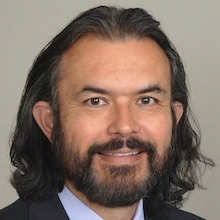 Professional headshot of a man with long, dark hair and a beard, wearing a suit and tie.