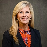 Professional headshot of a woman with long blonde hair, wearing a black blazer over a colorful patterned top.