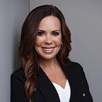 Professional headshot of a smiling woman with long, wavy brown hair, wearing a black blazer over a white top.
