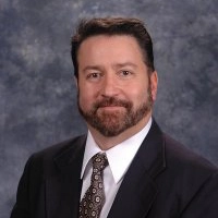 Professional headshot of a man with a beard, wearing a suit and tie against a gray background.